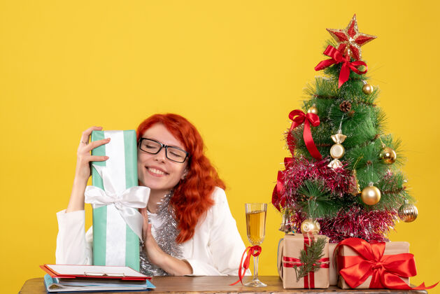 漂亮前视图：女医生拿着圣诞礼物和黄色背景上的圣诞树坐着医生礼物圣诞节