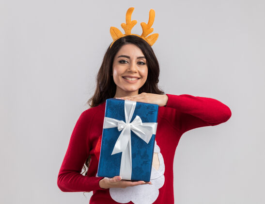 圣诞老人微笑的年轻漂亮女孩戴着驯鹿鹿角头带和圣诞老人毛衣拿着圣诞礼物包看拿着年轻年