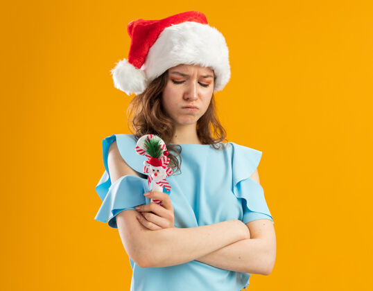 新的愤愤不平的年轻女子 穿着蓝色上衣 戴着圣诞帽 手里拿着圣诞糖果手杖 双手交叉 眉头紧锁蓝色交叉顶