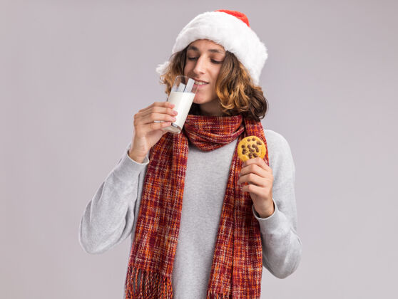 站着快乐的年轻人戴着圣诞老人的帽子 脖子上围着暖和的围巾 手里拿着饼干喝着牛奶围着温暖抱着