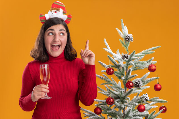 女孩穿着圣诞毛衣的年轻女孩戴着滑稽的头带 手里拿着一杯香槟 在橙色背景下的圣诞树旁开心又惊喜展示惊讶食指