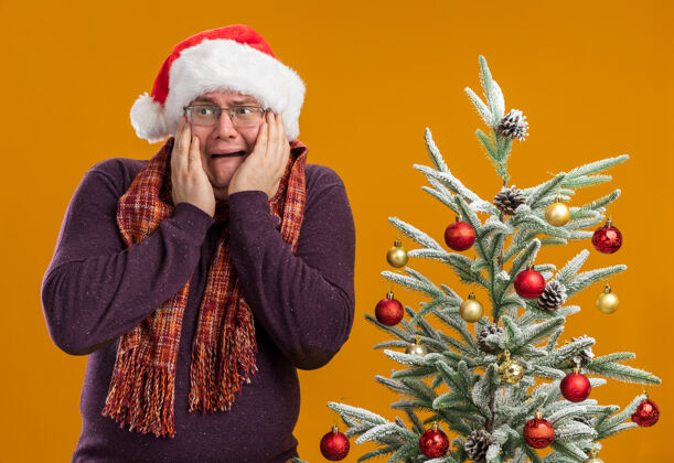 手戴着眼镜 戴着圣诞帽 脖子上围着围巾 站在装饰好的圣诞树旁 手放在脸上 看着橙色背景下孤立的一面装饰站着橙色