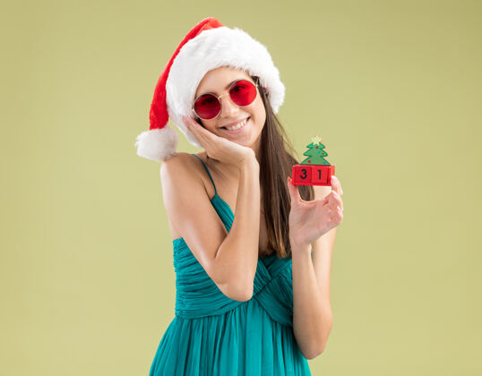 圣诞老人戴着太阳眼镜 戴着圣诞帽的年轻白种女孩高兴地把手放在脸上 手里拿着圣诞树饰物脸眼镜绿色