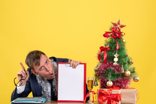 黄色商务人士拿着剪贴板和眼镜坐在圣诞树旁的桌子前 黄色的礼物树桌子眼镜