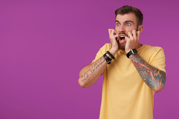男人带着纹身 留着短发 身穿黄色t恤的年轻胡须男子在紫色衣服上摆着姿势 惊恐地向一旁张望 双手捧着自己的脸身体手表手