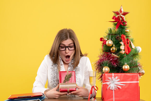 情感正面图：女医生坐在桌子前 拿着礼物 黄色背景上的圣诞树和礼盒电脑办公室树
