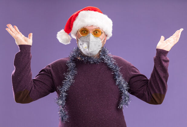 保护令人印象深刻的中年男子戴着圣诞帽 脖子上戴着金箔花环 戴着眼镜和防护面具 看着镜头帽子空面具