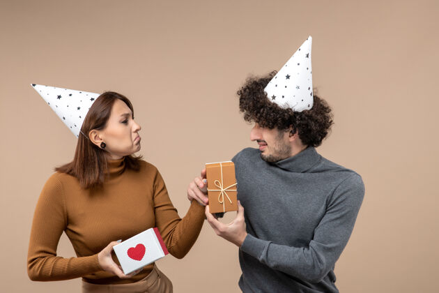 家伙新年拍摄与年轻夫妇戴新年帽紧张的女孩与心脏和困惑的家伙与礼物灰色肖像新的紧张