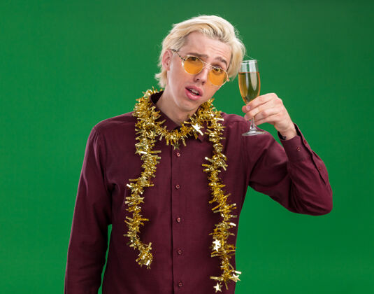 看可疑的年轻金发男子戴着眼镜 脖子上戴着金箔花环 头上拿着一杯香槟 看着隔离在绿色背景上的摄像机近眼镜戴着
