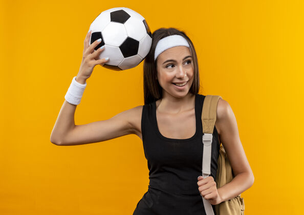 球快乐的年轻漂亮的运动女孩戴着头带和腕带和背袋把足球放在头上看着橙色空间上孤立的一面足球女孩漂亮