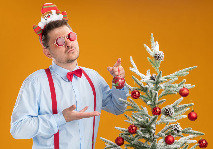 吊带戴着圣诞帽 戴着红眼镜 系着吊带领结的年轻人站在橙色墙上的圣诞树旁站着男人穿着