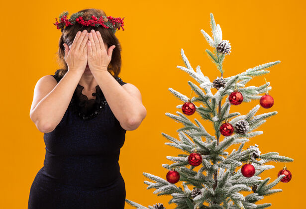 站着中年妇女戴着圣诞花环 脖子上戴着金属丝花环 站在装饰过的圣诞树旁 双手孤立地站在橙色的墙上 遮住脸脖子中年人圣诞节