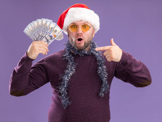 圣诞老人惊讶的中年男子戴着圣诞帽 脖子上戴着金箔花环 戴着眼镜 拿着并指着隔离在紫色墙上的钱惊喜周围金属片