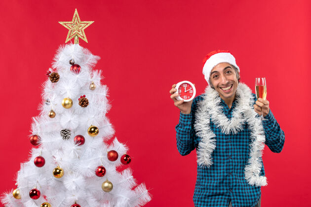 时钟快乐的年轻人戴着圣诞老人的帽子 手里拿着一杯葡萄酒 站在红地毯上的圣诞树旁的钟帽子礼物小伙子