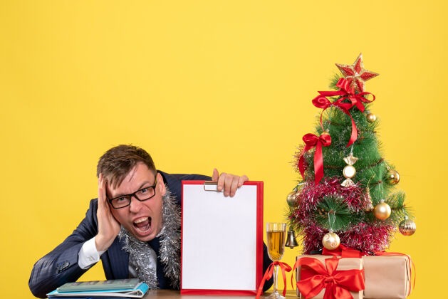 坐着前视图：手持剪贴板的商人坐在圣诞树旁的桌子旁 黄色的礼物丝带新郎桌子