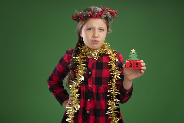 穿着小女孩戴着圣诞花环 穿着格子裙 脖子上挂着金属丝 手里拿着玩具立方体 表情严肃地写着圣诞节的日期站着看脖子