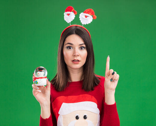 印象深刻年轻漂亮的白人女孩穿着圣诞老人的毛衣 头上戴着雪人的小雕像 孤零零地站在绿色的墙上毛衣年轻人雕像