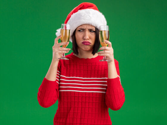 圣诞老人困惑的年轻女孩戴着圣诞帽拿着两杯香槟看着一个孤立的绿色墙壁上的复制空间穿着困惑香槟