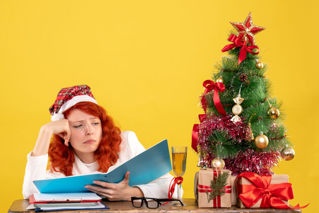 笔记本电脑正面图：女医生坐在桌子后面看黄色桌子上的文件 桌子上放着圣诞树和礼品盒办公室圣诞节女医生
