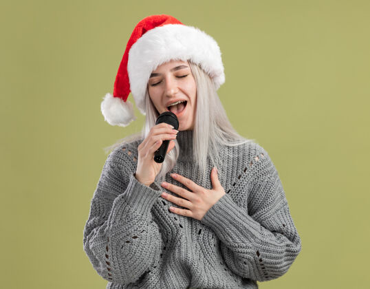 年年轻的金发女郎穿着冬衣 戴着圣诞帽 拿着麦克风 闭着眼睛快乐而积极地唱歌站着唱歌眼睛