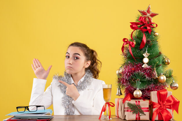 人正面图：女医生坐在黄色背景的桌子后面 还有圣诞树和礼品盒微笑插花坐着