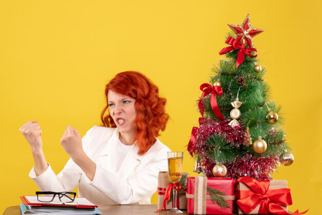 背景前视图：女医生坐在桌子后面 黄色背景上有圣诞礼物医生插花情绪