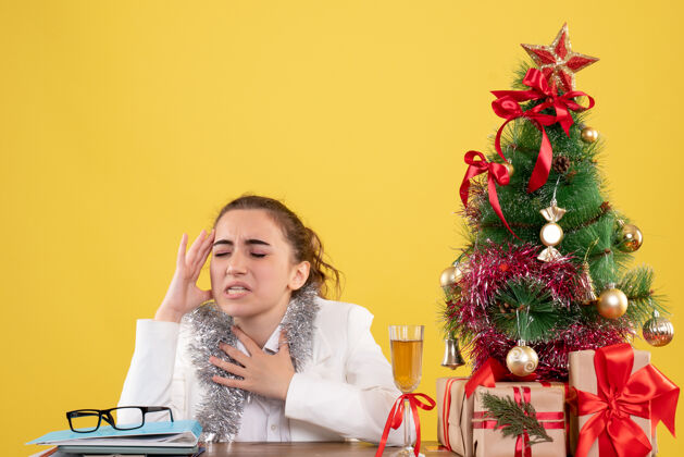 人正面图：坐在桌子后面的女医生头痛 背景是黄色的圣诞树和礼盒医生插花女医生