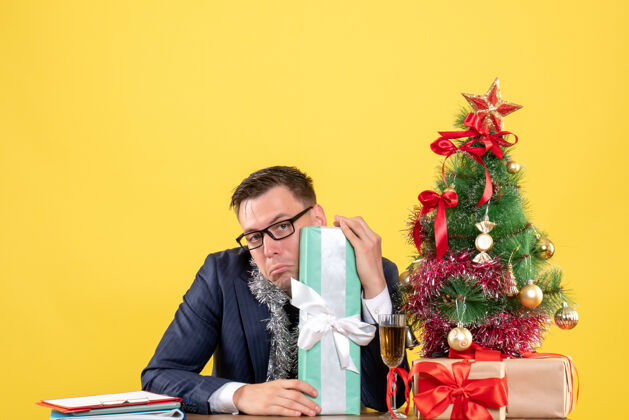 圣诞节沮丧的男人拿着礼物坐在圣诞树旁的桌子前 黄色的礼物黄色秘书沮丧