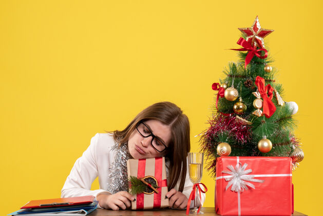 女性正面图：女医生坐在桌子前 拿着黄色背景的礼物 还有圣诞树和礼品盒情感女医生人
