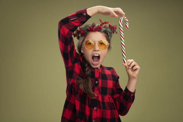 站着快乐而积极的小女孩戴着圣诞花环 穿着格子裙 手里拿着糖果手杖 开心地笑着衣服格子开心