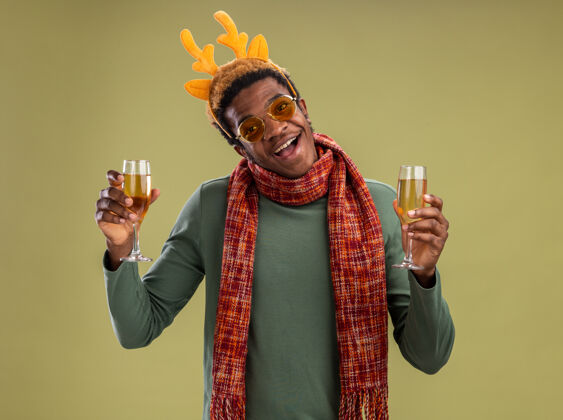 男人幸福的非洲裔美国人 戴着滑稽的边 戴着鹿角 脖子上围着围巾 手里拿着两杯香槟 站在绿色的墙上 开心地笑着围巾边缘鹿
