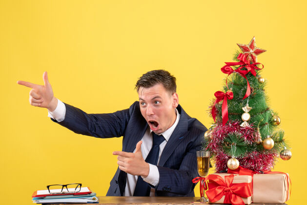 快乐前视图中的快乐男子手指的东西坐在圣诞树附近的桌子上 黄色的礼物什么树坐着