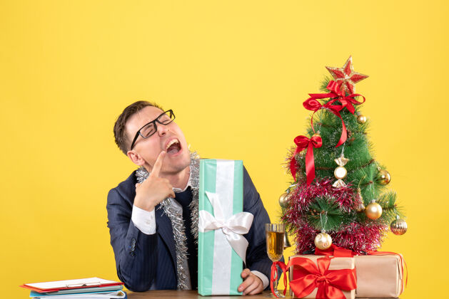 商人正面图：体贴的男人张嘴坐在圣诞树旁的桌子旁 黄色的礼物办公室桌子礼物
