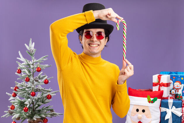 糖果身穿黄色高领毛衣 戴着黑色帽子 手持糖果手杖的快乐年轻人站在圣诞树旁 紫色的墙上挂着礼物手杖穿着站着