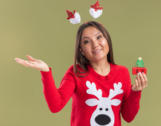 圣诞节高兴的歪着头的年轻亚洲女孩戴着圣诞发箍 手拿着圣诞玩具点 在橄榄绿的墙上孤立着头年轻边