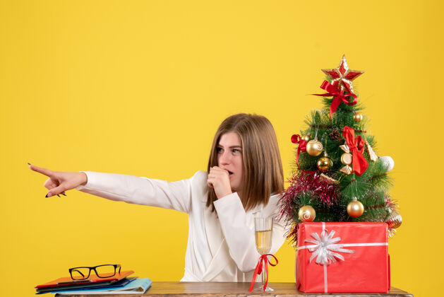 女性前视图：女医生坐在黄色地板上的桌子前 手里拿着圣诞树和礼品盒坐着人圣诞节