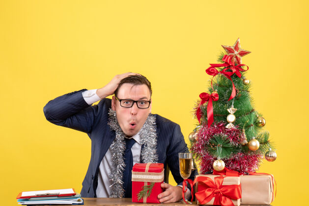 黄色困惑的男人抱着他的头坐在圣诞树旁的桌子前 黄色的礼物人新郎圣诞