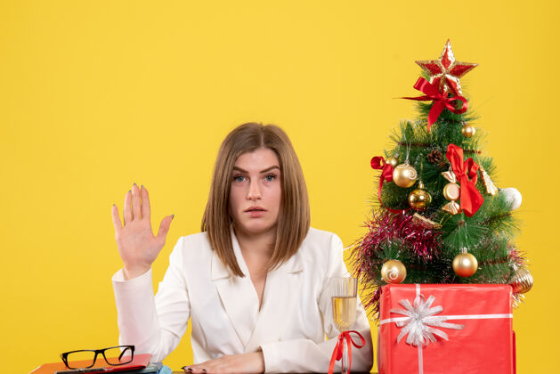 圣诞节正面图：女医生坐在黄色背景的桌子前 放着圣诞树和礼品盒背景人微笑