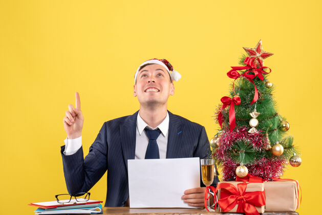 商人正面图是兴高采烈的男人坐在圣诞树旁的桌子上看黄色的礼物人看黄色