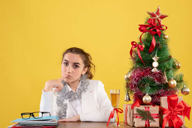 微笑前视图：女医生坐在桌子后面 在黄色背景上思考着圣诞树和礼品盒女医生快乐人
