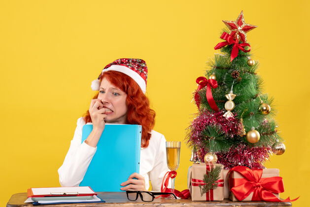 微笑正面图：女医生坐在桌子后面 拿着黄色背景的文件 还有圣诞树和礼品盒快乐女性微笑