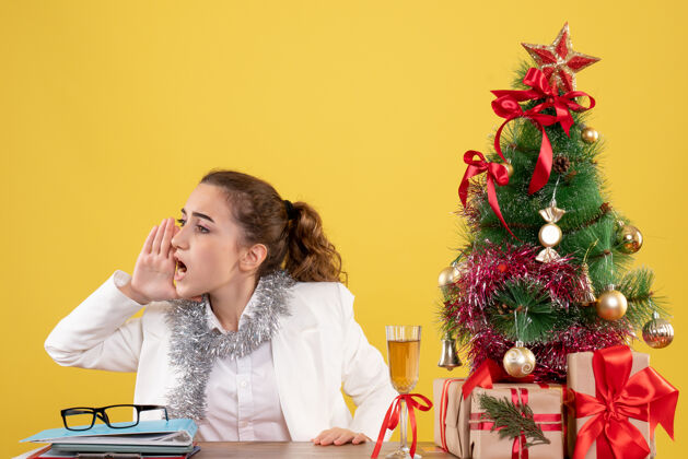 呼叫前视图：女医生坐在桌子后面 用圣诞树和礼品盒在黄色背景上呼唤某人新郎快乐微笑