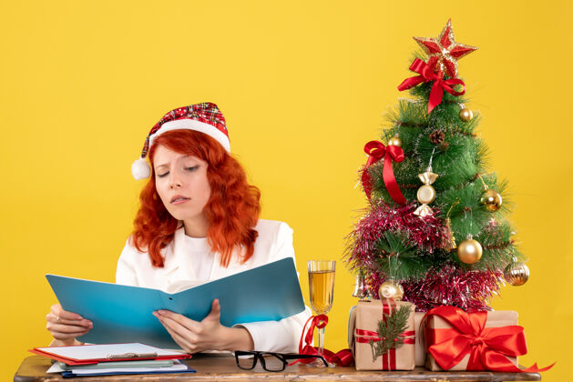 抱着前视图女医生坐在桌子后面 拿着黄色背景上的文件和圣诞树年份礼物背景