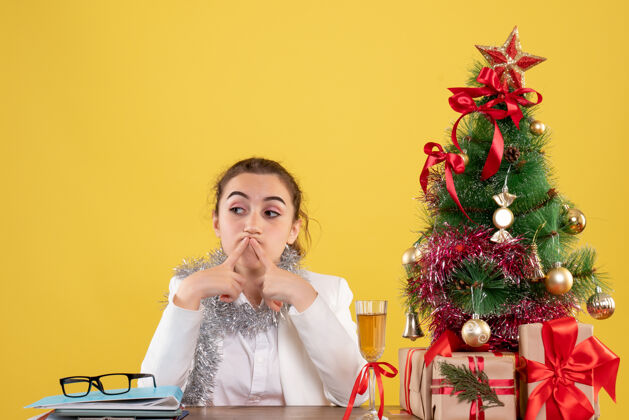 健康正面图：女医生坐在桌子后面 黄色背景上无聊地摆放着圣诞树和礼品盒女医生肖像女性
