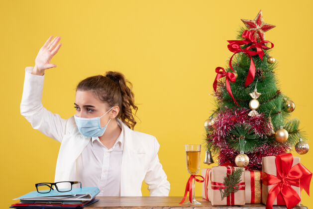 人正面图女医生戴着黄色背景的无菌口罩坐在圣诞树和礼盒里微笑成人无菌