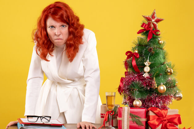 新的正面图：女医生坐在桌子后面 拿着黄色背景上愤怒的圣诞礼物 还有圣诞树和礼盒前面成人背景