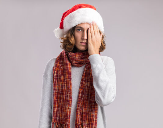 手年轻人戴着圣诞老人帽 脖子上围着暖和的围巾 严肃的脸遮住一只眼睛 手站在白色的背景上看着摄像机看相机盖