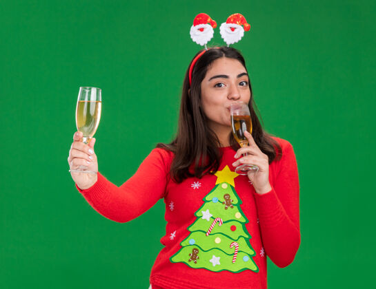 年戴着圣诞老人头巾的年轻白人女孩高兴地捧着香槟 喝着香槟 香槟酒被隔离在绿色背景上 并留有复印空间年轻圣诞快乐圣诞节
