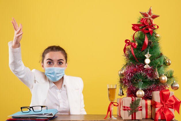 外套正面图女医生戴着防护面具坐在黄色背景上与圣诞树和礼品盒争吵健康新郎医生