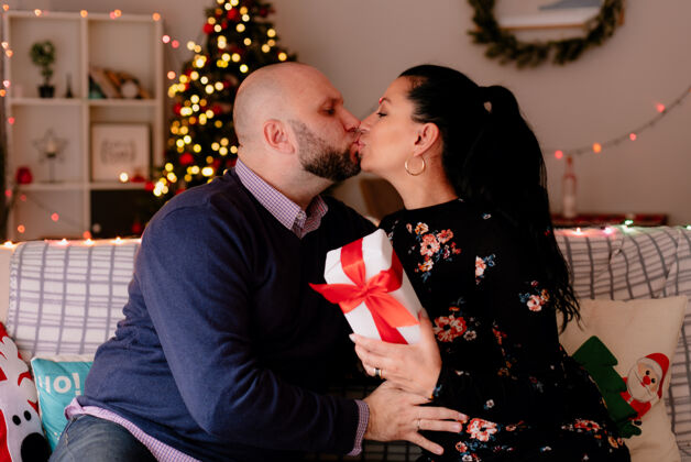 包裹爱浪漫的夫妻在家过圣诞节坐在客厅沙发上妻子拿着礼包亲吻嘴唇生活嘴唇抱着